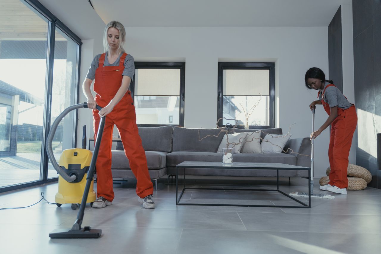 about-us Two women in red overalls vacuuming and sweeping a modern living room.